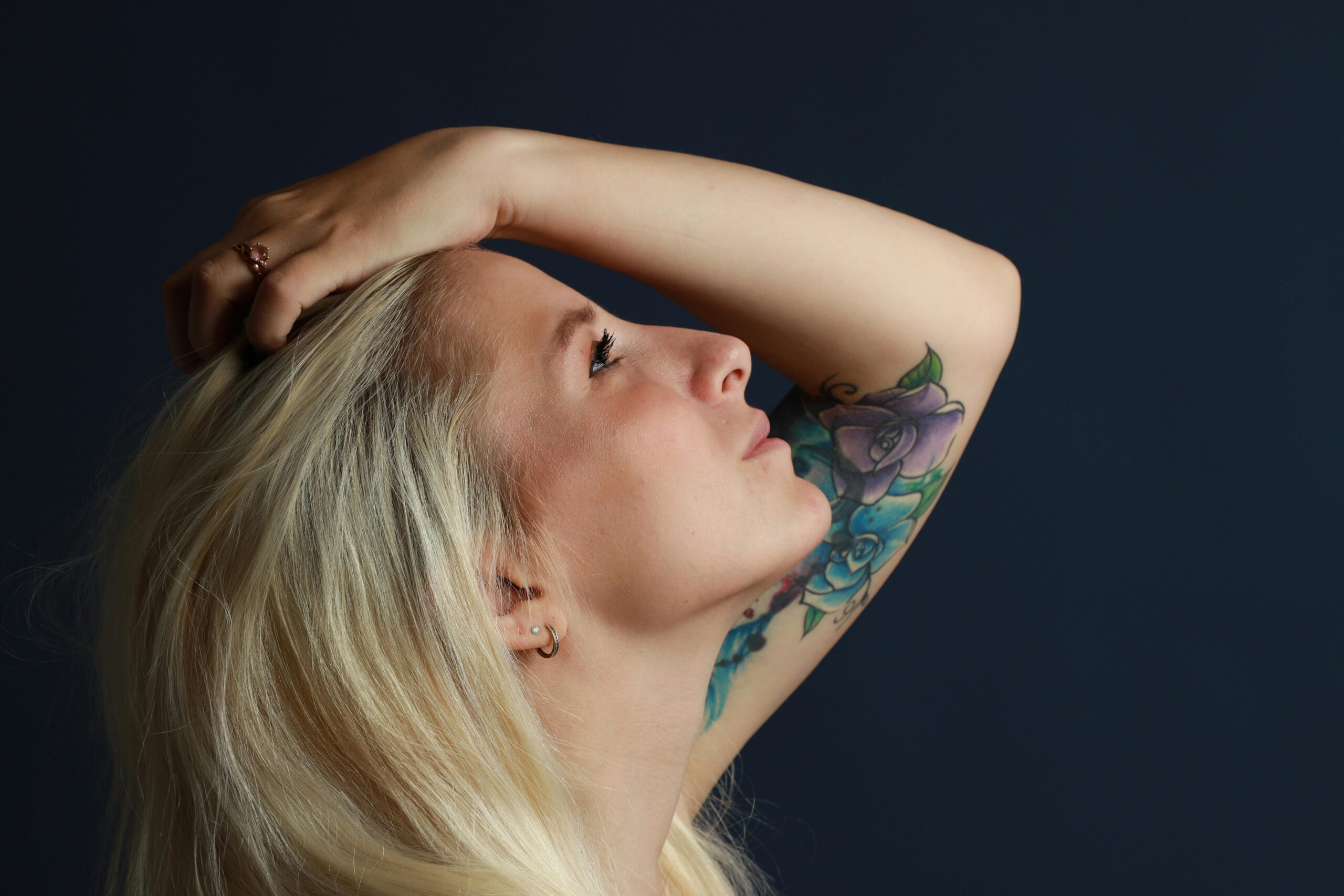 Profile portrait of a blonde woman with a floral tattoo on a dark background.